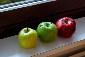Red, green and yellow apples lie on the windowsill by the window.