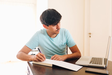 young man with laptop writing down in a notebook