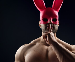 Brutal sweaty strong young man with naked pumped up upper body in pink rabbit mask standing showing silence sign with fingers over grey background. Sport men body and sexual games concept