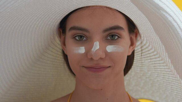 Close-up beauty portrait of young good-looking brown-haired caucasian woman in a big white hat with sun cream on her cheeks and nose blows a kiss for the camera | Face skin protection concept