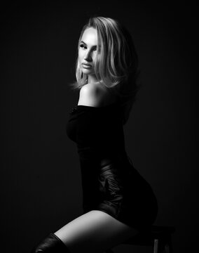 Young pretty sexy blonde woman in black clothes with open shouplder and high boots sitting on stool looking at camera over dark background. Fashion, style, sexy wear for women concept