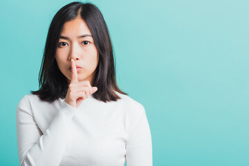 Young beautiful Asian woman holding index finger on her mouth lips, Portrait female hush silence, studio shot isolated on a blue background, Gesture of shhh