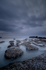 Obraz premium Dreamy seascape for background. Beach with rocks and stones. Low tide. Blurred foggy water. Cloudy sky. Slow shutter speed. Soft focus. Copy space. Mengening beach, Bali