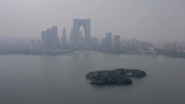 Aerial panning shot of green island at Jinji Lake against buildings, drone flying near cityscape against sky - Suzhou, China