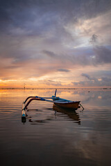 Seascape. Fisherman boat. Traditional Balinese boat jukung. Fishing boat at the beach during sunrise. Water reflection. Cloudy sky. Sunlight on horizon. Slow shutter speed. Bali