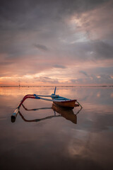Seascape. Fisherman boat. Traditional Balinese boat jukung. Fishing boat at the beach during sunrise. Water reflection. Cloudy sky. Sunlight on horizon. Slow shutter speed. Bali