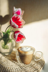Fototapeta na wymiar cup of coffee with lush milk foam and three blooming tulips in glass vase on table with woven boho napkin in hard natural light with shadows. vertical cozy cute spring breakfast, selective focus