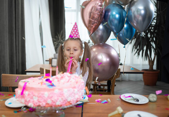 a little girl celebrates her birthday, stands alone near the cake and blows a festive pipe. Air balloons.