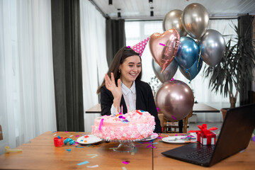 portrait of a young beautiful woman sitting at a table and celebrating her birthday. Communicate via webcam and laugh merrily. Waving hand a sign of greeting.