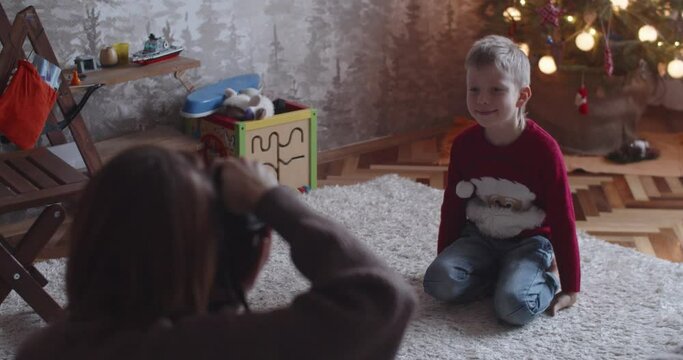Faceless photographer shooting cheerful kid boy at home interior on new year eve. Mother making photo of son child sitting by Christmas tree laughing slow motion. Happy holiday memories concept