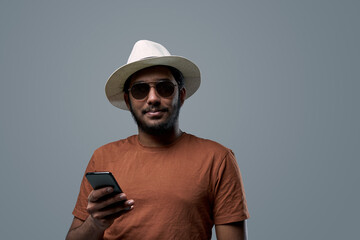 Self confident and bearded indian guy in orange shirt poses in gray background holding his smartphone.