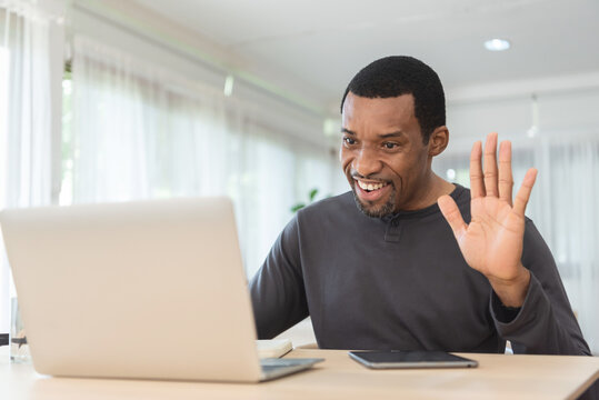 African American Freelancer waving hand while having video call with friends or family on laptop at home office. Black Male making online meeting and working on notebook.