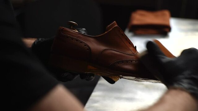 Close-up view of professional shoemaker in black gloves polishing light brown leather shoes with brush during restoration working. Concept of footwear maintenance captured. Shooting in slow motion.