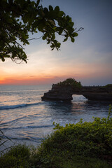 Fototapeta na wymiar Natural arch. Batu Bolong temple on the rock during sunset. Seascape background. Motion waves. Foreground with tree branches. Copy space. Vertical layout. Tanah Lot, Bali