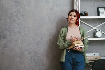 Thinking woman with red hair writing and holding books while standing