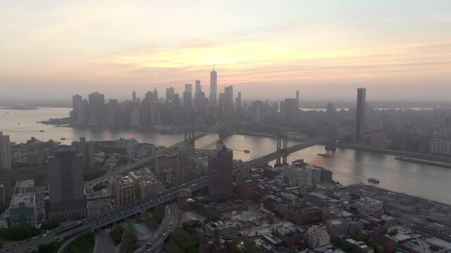 Aerial shot of vehicles and buildings in city by East River, drone flying forward towards Manhattan against sky during sunset - New York City, New York