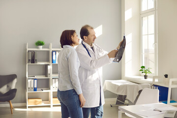 Mature doctor at clinic showing MRI or X-ray scan to patient. Young woman looking at CT image and listening to medical specialist's explanation. Early diagnosis of cancer, treatment of physical injury