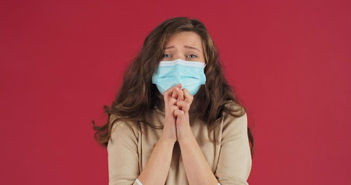 Young girl with curly hair wears medical protective mask spins fingers praying hoping for success asks fate and god for good test results for coronavirus stands isolated on red background in studio