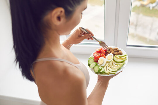 Young brunette fitness woman athete in sportswear standing and eating healthy meal with eggs, nuts and vegetables before or after workout at home, top view. Active healthy lifestyle, clean eating