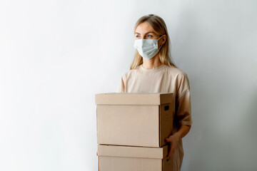 A European white woman wearing a protective medical mask on her face is standing against a wall holding large cardboard boxes. Delivery of online orders. New normal life into an epidemic.