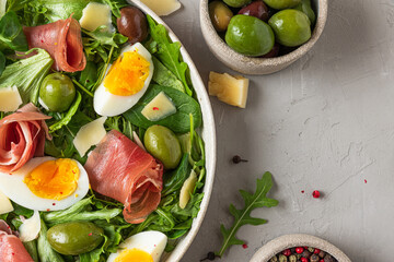 Prosciutto di Parma salad with olives, parmesan cheese, eggs and arugula on gray background. Italian appetizer. Top view