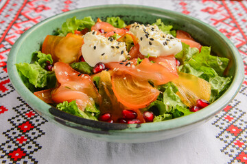 Delicious healthy salmon salad with cheese and pomegranate seeds served in a bowl over traditional Ukrainian tablecloth.