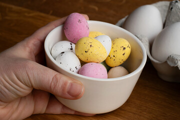 Hand holding up a bowl filled with chocolate Easter eggs in various colors. In the background is an egg carton on wooden table.