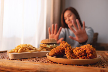 A woman making hand sign to refuse a hamburger, french fries and fried chicken on the table for...