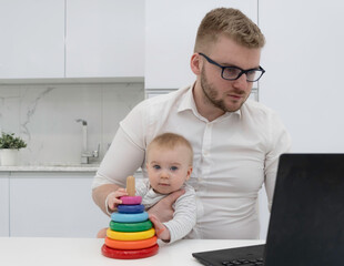 Handsome bearded blond man wearing white shirt working in the kithen, at home with laptop, holding cute baby play with pyramid. Work from home with kids concept