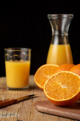 Close-up of half orange, bottle, knife, out of focus oranges on wooden table, black background, vertical, with copy space