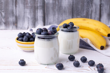 A healthy breakfast of Greek yogurt in glass jars and fresh blueberries and ripe bananas at a rustic white table. Selective focus on glass jar.
