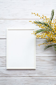 White blank photo frame with a bouquet of mimosa on a white wooden table. Minimalistic, mockup. view from above