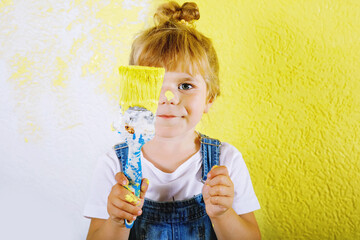 Cute little toddler girl painting the wall with yellow color in new house. Family repair apartment home. Happy child girl paints the wall, choosing color with palette, having fun with brush, indoors