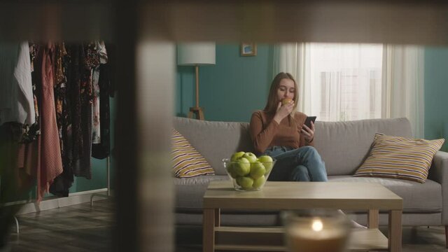 Pretty woman in blue jeans and brown sweater sits on beige sofa on window background, bit red apple and choked. Trying to cough. On table is glass jar with green apples. Camera dollies horizontally