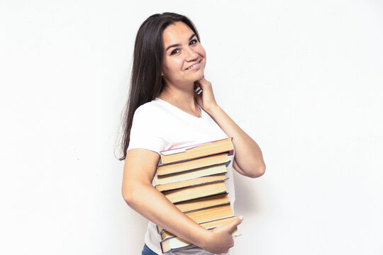Young smiling student woman in white t-shirt holding textbooks and books in a pile on a white backdrop. College education concept. Positive emotions, facial expression