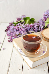 Romantic background with a cup of tea, lilac flowers and a book over a white wooden table. Leisure concept, spring breakfast
