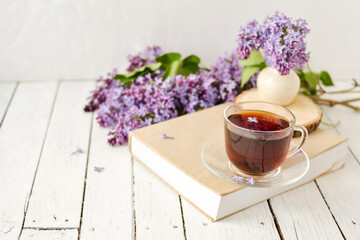 Romantic background with a cup of tea, lilac flowers and a book over a white wooden table. Leisure concept, spring breakfast