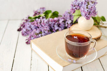 Romantic background with a cup of tea, lilac flowers and a book over a white wooden table. Leisure concept, spring breakfast
