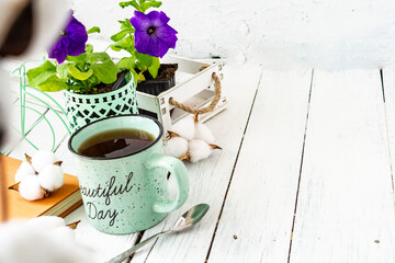 Close-up of a cup of tea on a wooden white table with blurred background, front blur. Still life with flower, book, teaspoon, cotton box. Spring breakfast. Copy space