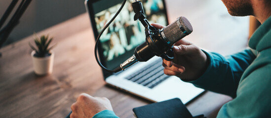 A man host streaming his audio podcast using microphone and laptop at his small broadcast studio, close-up - obrazy, fototapety, plakaty