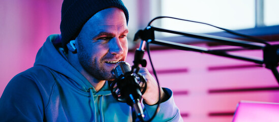Trendy podcast creator streaming live audio broadcast at his home studio using stylish cyber punk...