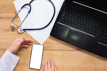 Cropped shot top view of doctor hands using smartphone mockup on medical office desk. Blank screen mobile phone for graphic display montage near laptop computer