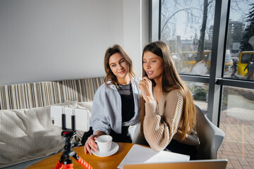 Fototapeta premium Two young beautiful girls are sitting in a cafe, recording video blogs and communicating on social networks.