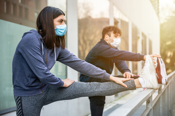 Young couple outdoors in the city do stretching training exercises with mask to protect themselves from Coronavirus Covid-19 infections - Man and woman train for the marathon and have fun together