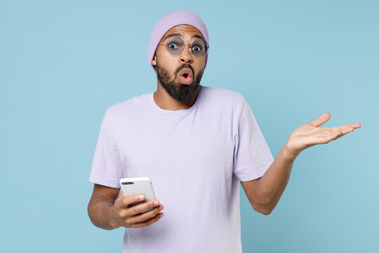 Young shocked indignant unshaven student black african man 20s in violet t-shirt purple hat glasses using mobile cell phone spread hand oops gesture isolated on pastel blue background studio portrait.