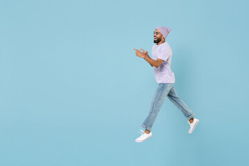 Fototapeta na wymiar Full length young cool smiling happy fun black african man 20s wear violet t-shirt hat glasses point index finger aside on workspace area jump high isolated on pastel blue background studio portrait.