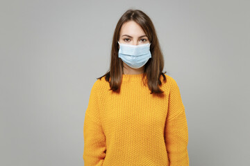 Young pretty woman 20s wearing casual knitted yellow sweater in sterile face mask to safe from coronavirus virus covid-19 during pandemic quarantine isolated on grey color background studio portrait