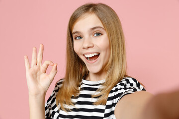 Close up little blonde smiling happy kid girl 12-13 years old in striped oversized t-shirt do selfie shot on mobile phone show ok okay gesture isolated on pink background children Childhood concept.