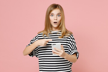 Little blonde shocked kid girl 12-13 years old wearing striped oversized t-shirt point index finger on mobile cell phone chat isolated on pastel pink background children. Childhood lifestyle concept.