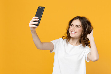 Naklejka premium Young smiling happy friendly positive caucasian woman 20s in white basic casual t-shirt do selfie shot on mobile phone show thumb up like gesture isolated on yellow orange background studio portrait.
